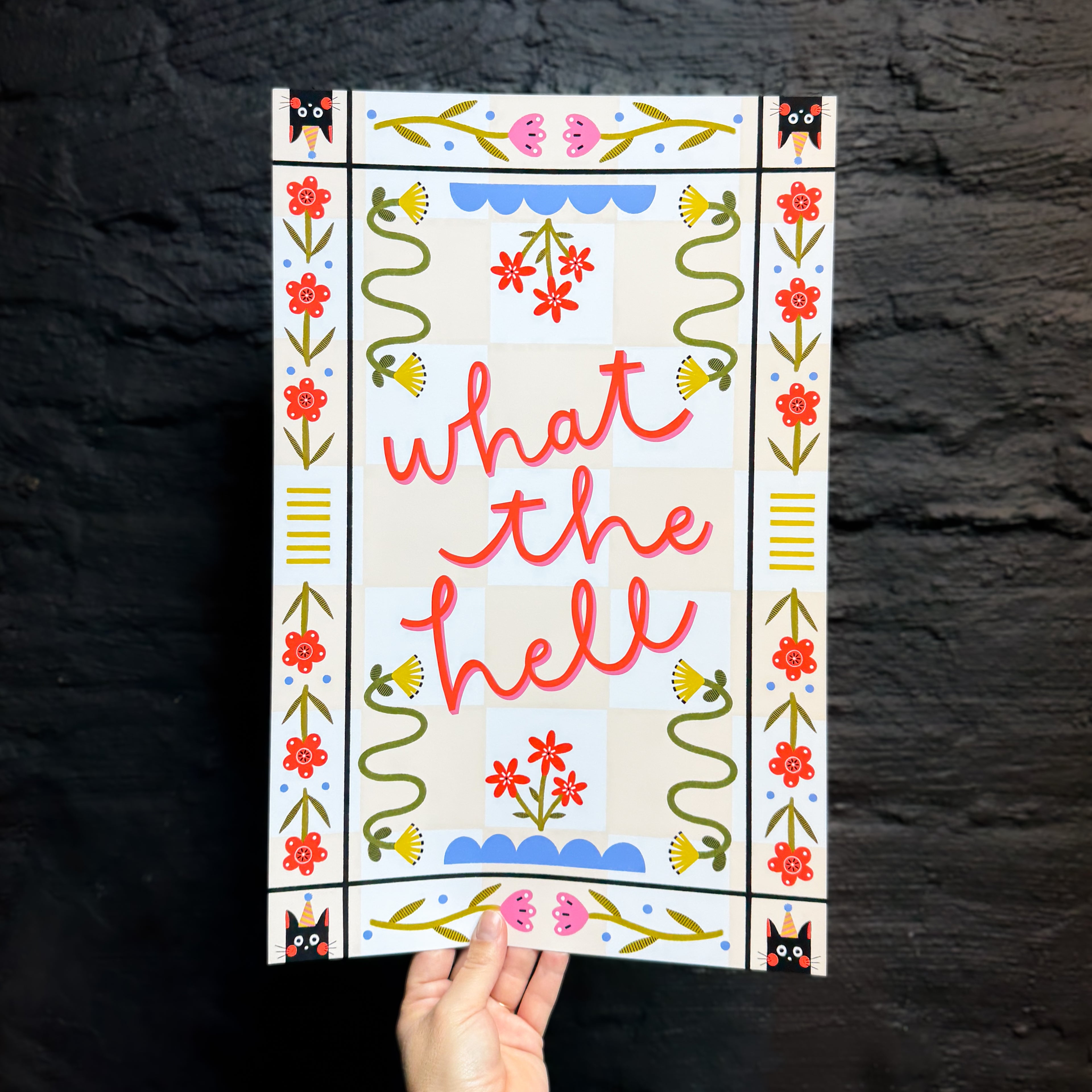 A hand holding an 11x17 art print with a peach checkered background that says 'what the hell' in the middle, surrounded by folk art style flowers and cats with party hats in the corners.