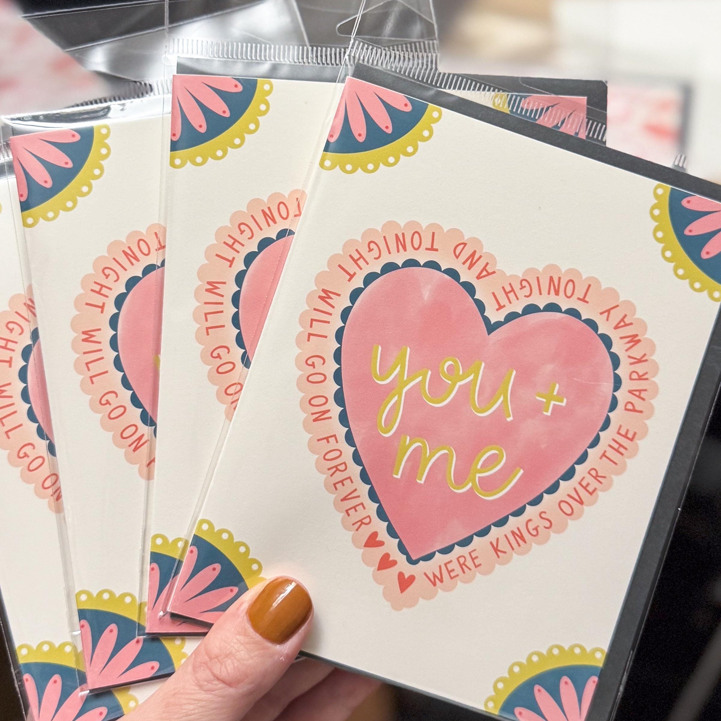 A  hand holding greeting cards with a heart in the middle that says 'you and me were kings over the parkway tonight and tonight will go on forever' with three small hearts to fill space. All four corners are embellished with a scalloped flower.