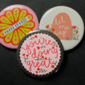 A round pinback button featuring a light pink background with a grey/green scalloped edge and red flowers with the text 'you're doing great'. The pin is surrounded by other pin designs.