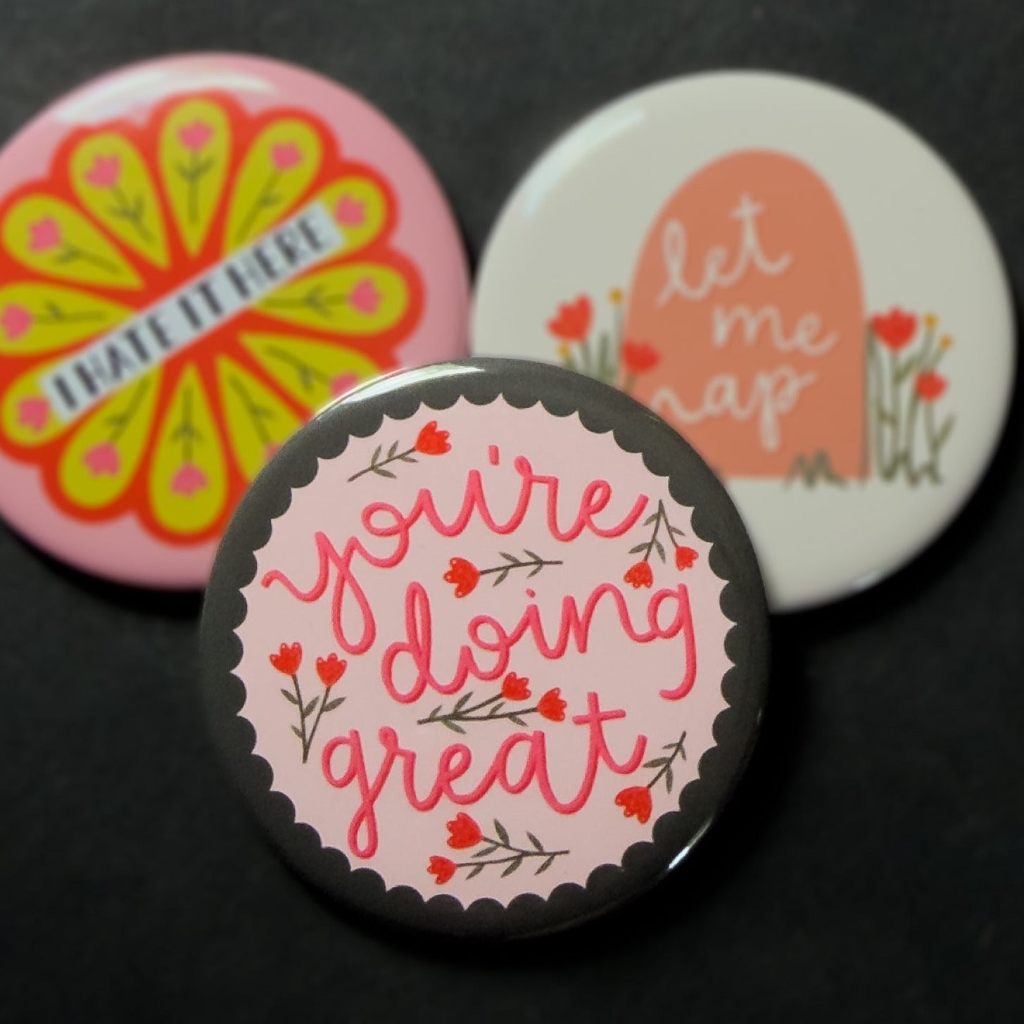 A round pinback button featuring a light pink background with a grey/green scalloped edge and red flowers with the text 'you're doing great'. The pin is surrounded by other pin designs.