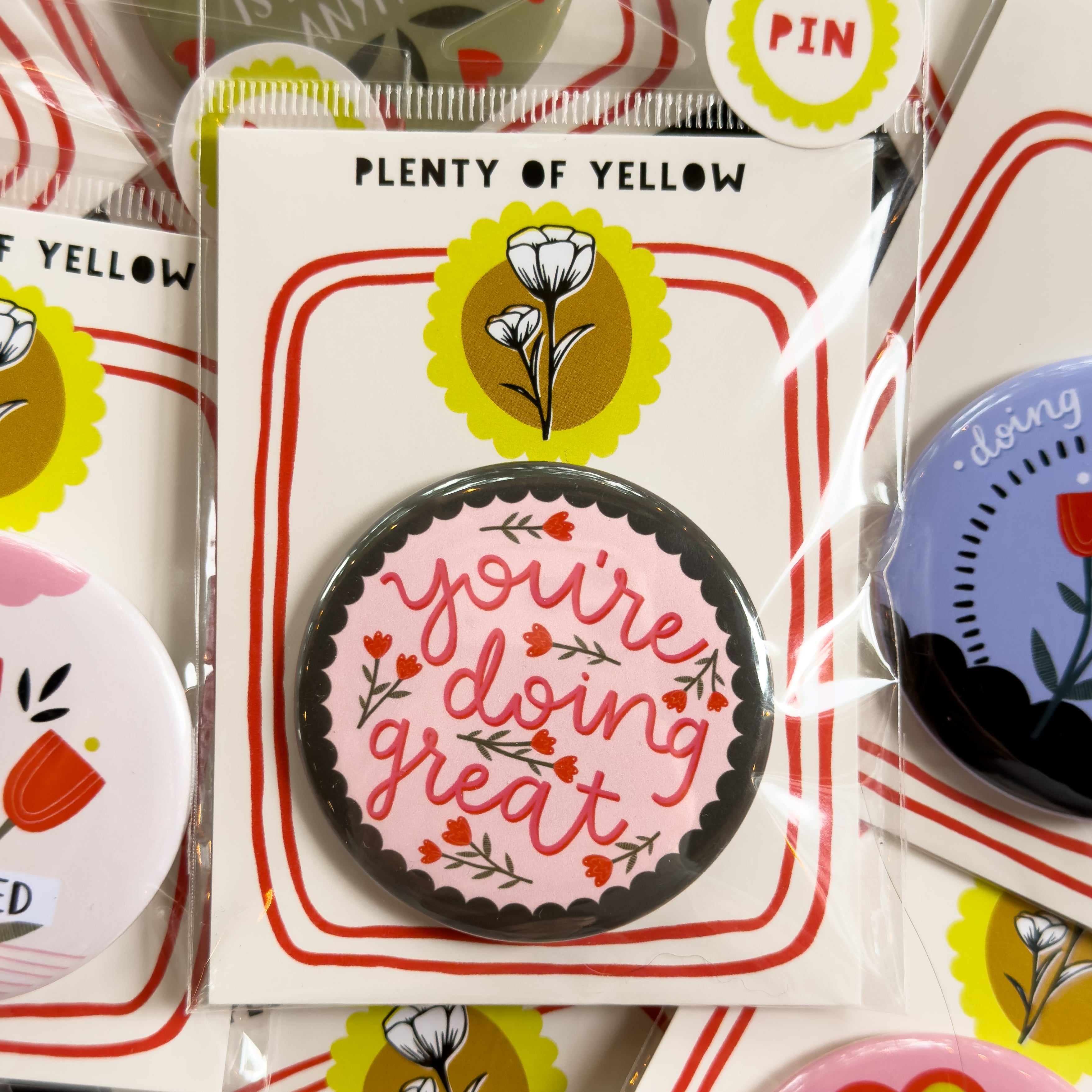 A round pinback button in clear plastic packaging, featuring a light pink background with a grey/green scalloped edge and red flowers with the text 'you're doing great'. The pin is mounted on a white backing card with red borders and a yellow logo, under the brand name 'PLENTY OF YELLOW'.
